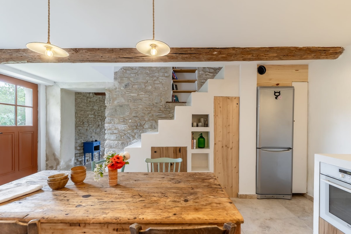 A rustic kitchen features a large wooden dining table adorned with a vase of flowers. Exposed wooden beams and stone wall accents add charm. A modern refrigerator and a white microwave are visible. The staircase leading to an upper level offers functional storage spaces.