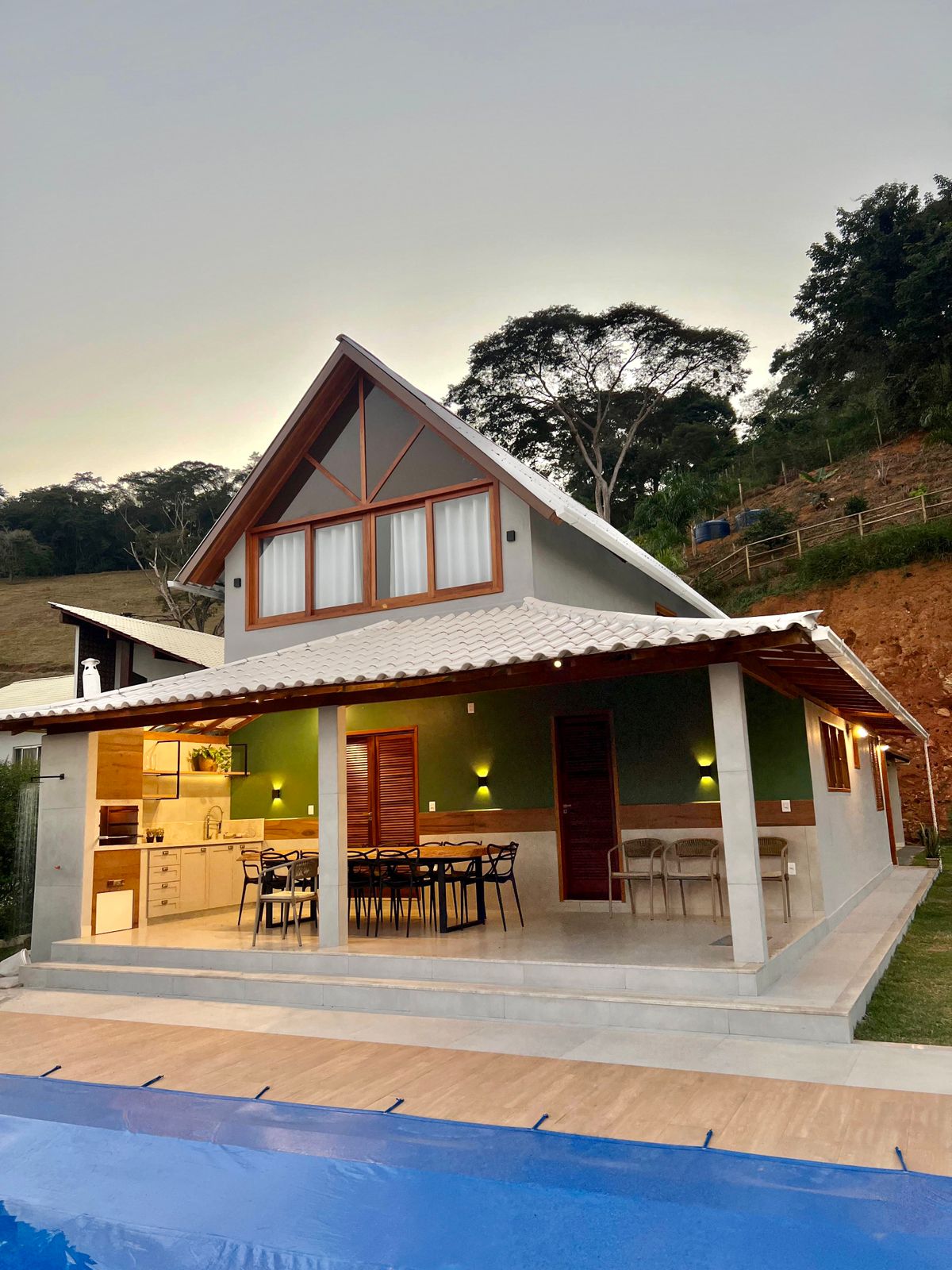 A charming two-story chalet is presented, showcasing a sloped roof and inviting outdoor patio. The exterior features wooden accents, large windows, and a well-maintained garden area, while a sparkling pool extends along the side, creating a serene setting. Warm lighting enhances the inviting feel of the space.