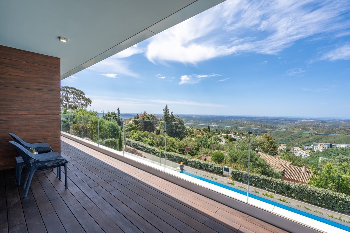 A spacious terrace features a wooden deck and modern glass railing, providing unobstructed views of the surrounding landscape. Two blue chairs are positioned for relaxation, inviting guests to enjoy the expansive horizon under a clear sky.