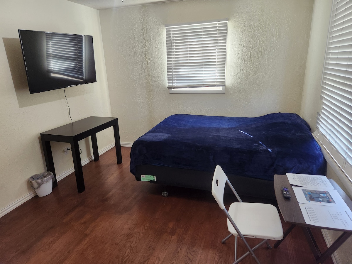 A cozy room features a spacious bed covered with a dark blue throw. A wall-mounted television is positioned across from a small table and folding chair. Natural light enters through a window with blinds, enhancing the warm ambiance of the wooden floor.