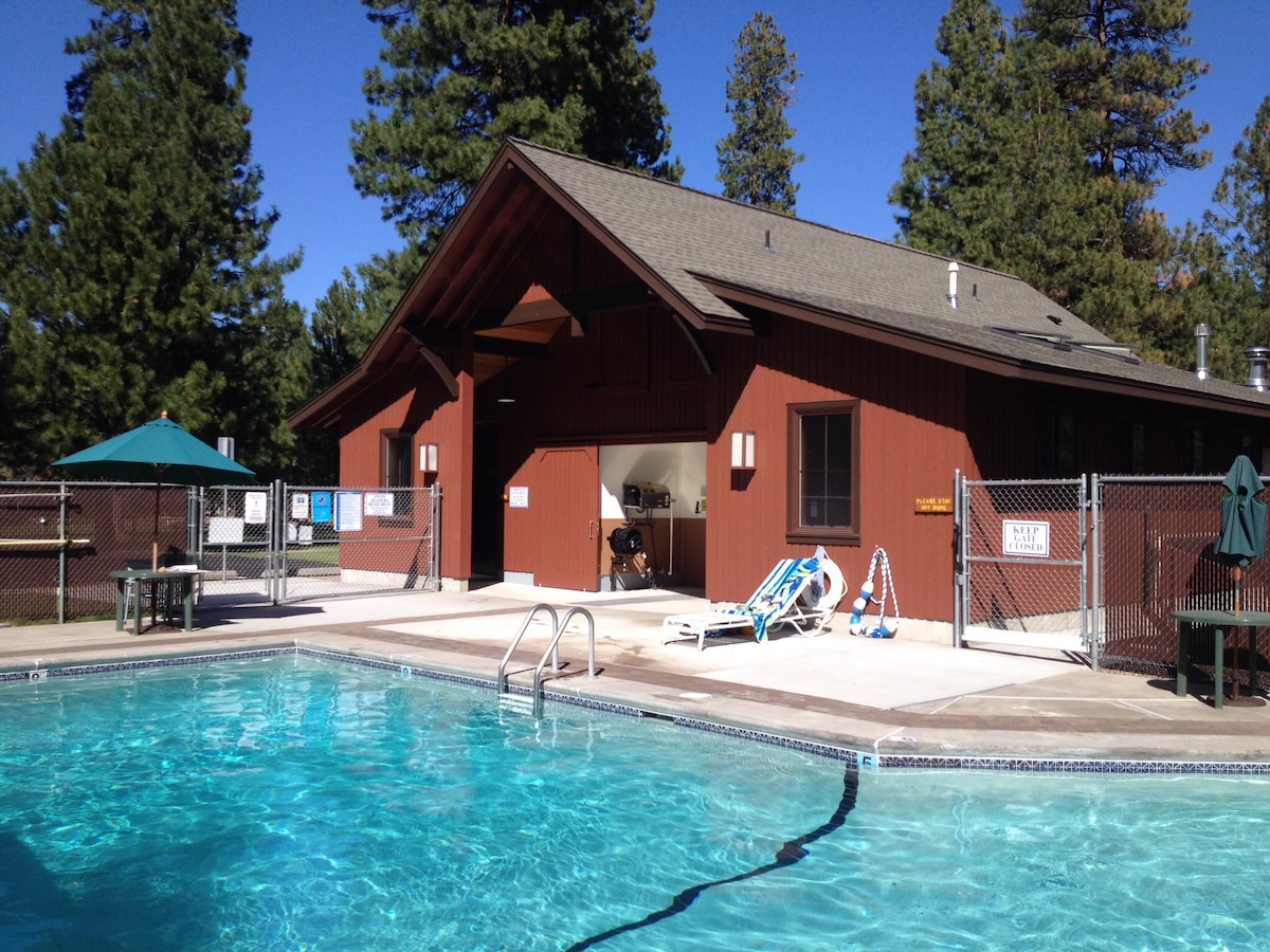 A seasonal pool area features clear water inviting for a swim. Surrounding the pool are lounge chairs and shaded areas beneath umbrellas. A large building with a rustic design stands nearby, offering amenities and shelter for guests.