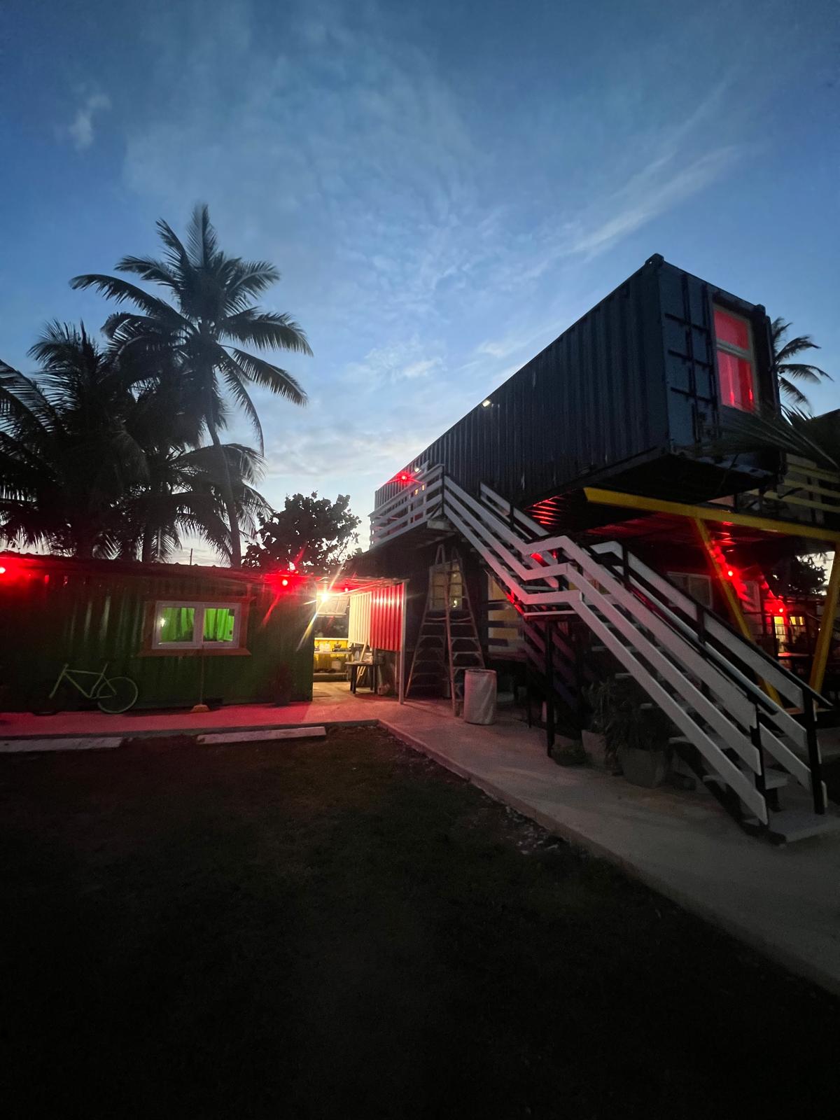 A unique shipping container structure is surrounded by palm trees, illuminated by soft red lights. A set of stairs leads to an upper level, while a ground-level unit is visible. The evening sky features soft clouds, creating a calm atmosphere.