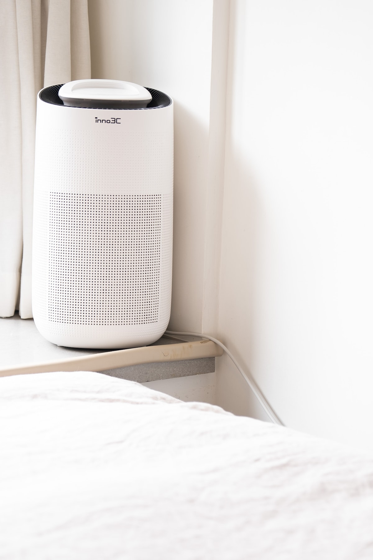 An air purifier stands quietly in the corner of a bedroom, placed beside a fluffy white bed covering. Soft natural light filters through nearby curtains, illuminating the minimalist design of the white purifier against the pale wall.