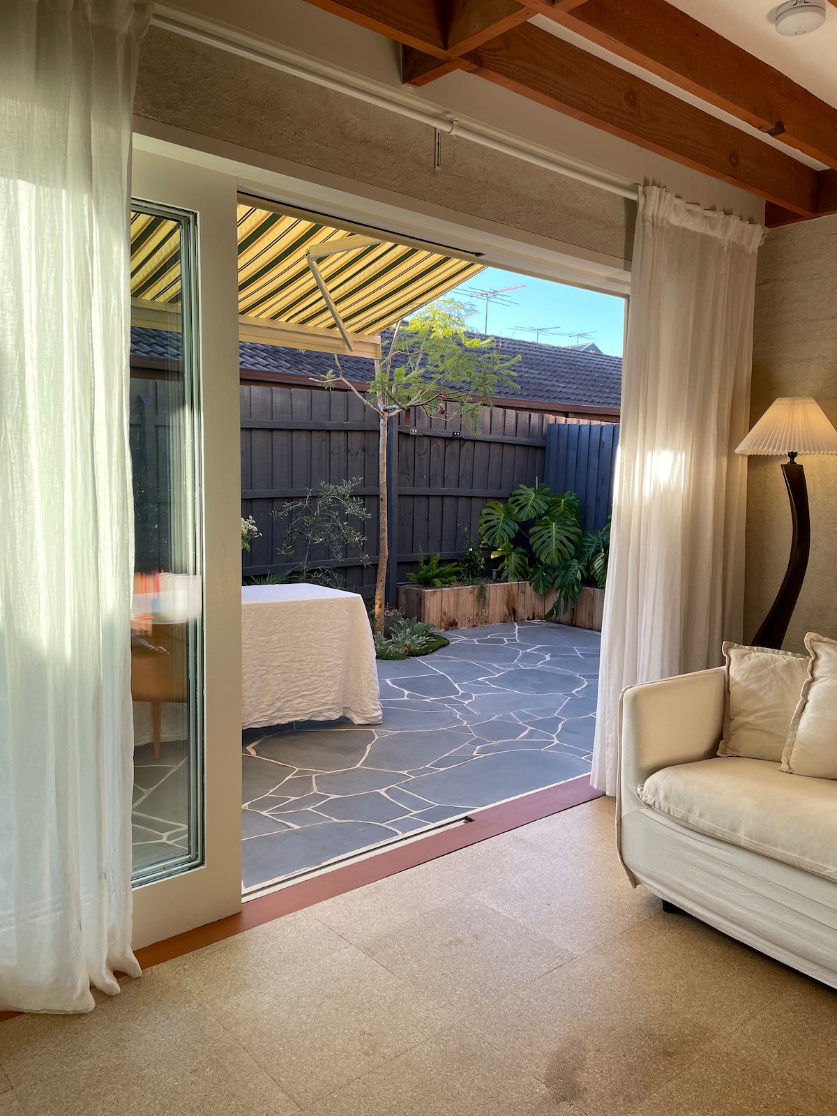 Natural light flows into the living space through glass doors, revealing a tranquil courtyard with a stone-patterned floor. A table is set under a striped awning, surrounded by green plants. Light curtains gently frame the entrance, enhancing the inviting ambiance.