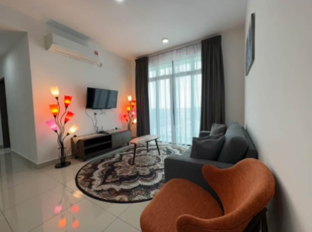 A cozy living area features a modern couch and a stylish orange accent chair. A wooden coffee table stands on a decorative rug, while floor lamps with colorful shades provide ambient lighting. Large glass doors lead to a view beyond, framed by dark curtains.