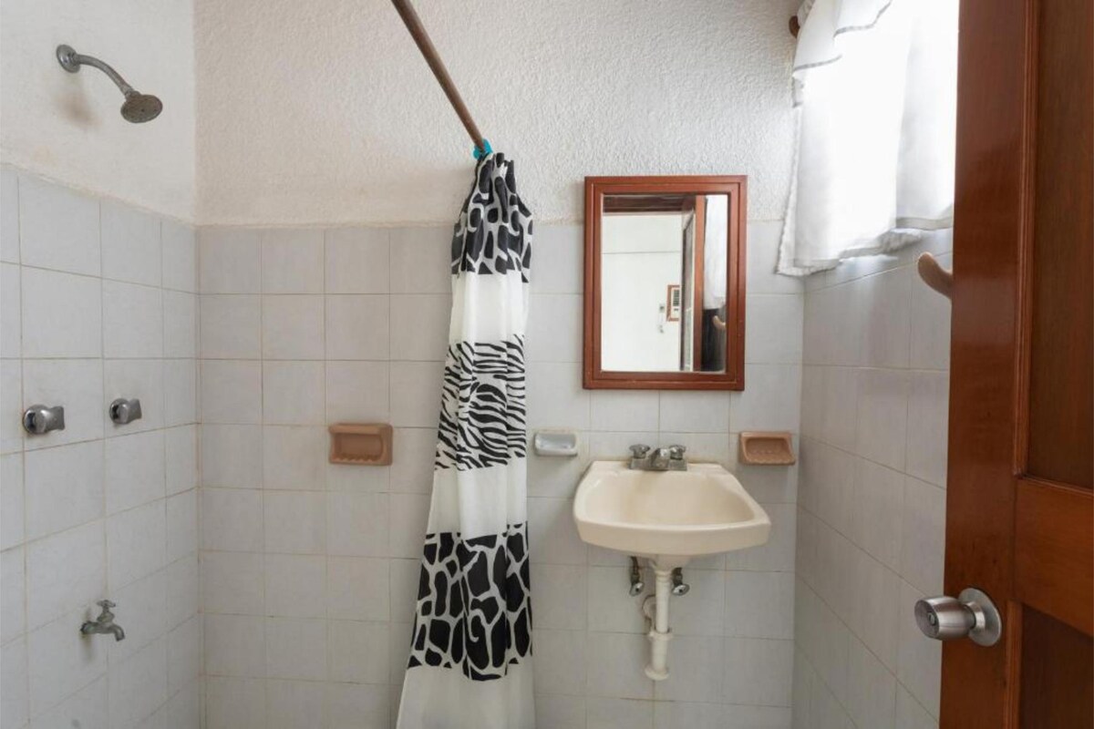 A clean bathroom features white tiled walls and a small sink beneath a mirror. A shower area is enclosed, with a black and white striped curtain providing privacy. Fixtures are visible, alongside wall-mounted holders for toiletries.
