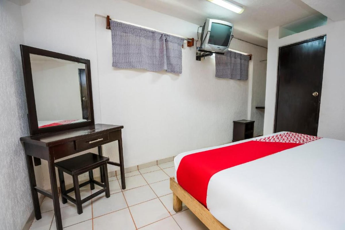 A clean and functional room features a spacious bed dressed in crisp white linens with a red accent. A wooden desk with a matching stool is positioned nearby, complemented by a wall-mounted television. Natural light filters through curtains at the window, illuminating the neutral walls.