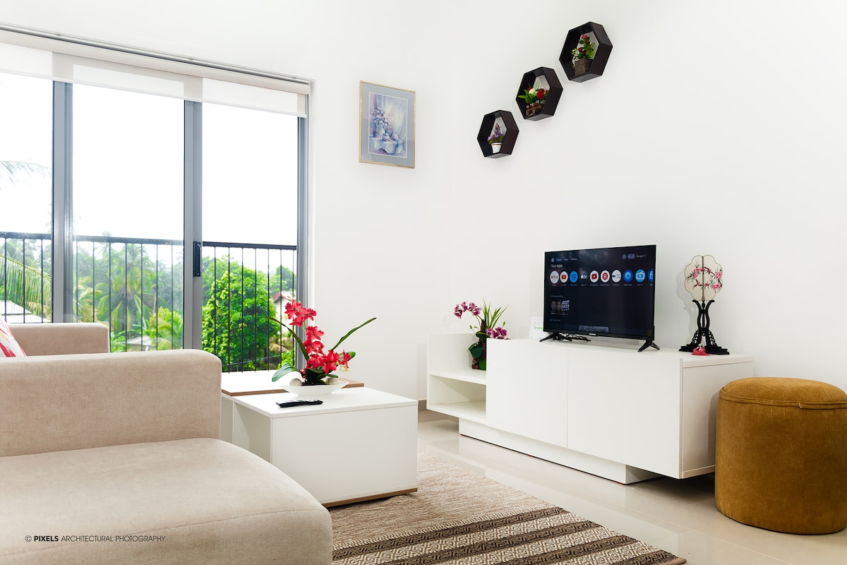 A bright living area features a comfortable white sofa positioned opposite a TV unit. Decorative plants and a wall-mounted display enhance the space. Natural light filters through large windows, showcasing views of the greenery outside. A cozy ottoman adds an additional seating option.