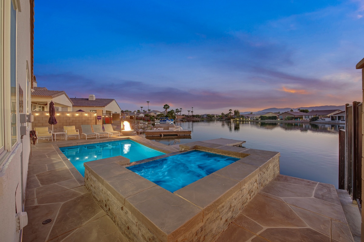 A serene outdoor area is highlighted by a pool and spa, both featuring illuminated blue water. The surrounding stone deck offers lounge chairs, while a tranquil lake view extends in the background, reflecting the colorful sky.