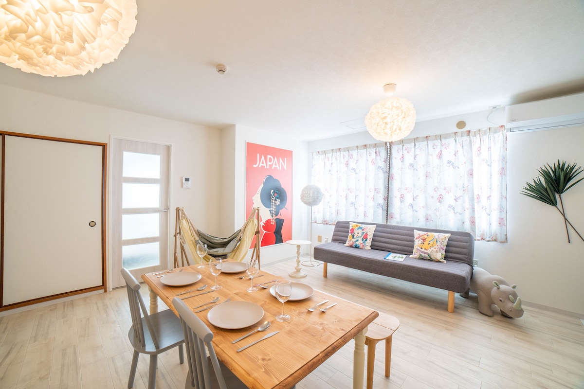 A bright dining area features a wooden table set for a meal, accompanied by light-colored chairs. A plush gray sofa adorned with colorful cushions is positioned nearby. Natural light filters through floral-patterned curtains, enhancing the inviting space. A hammock adds a unique touch to the room.