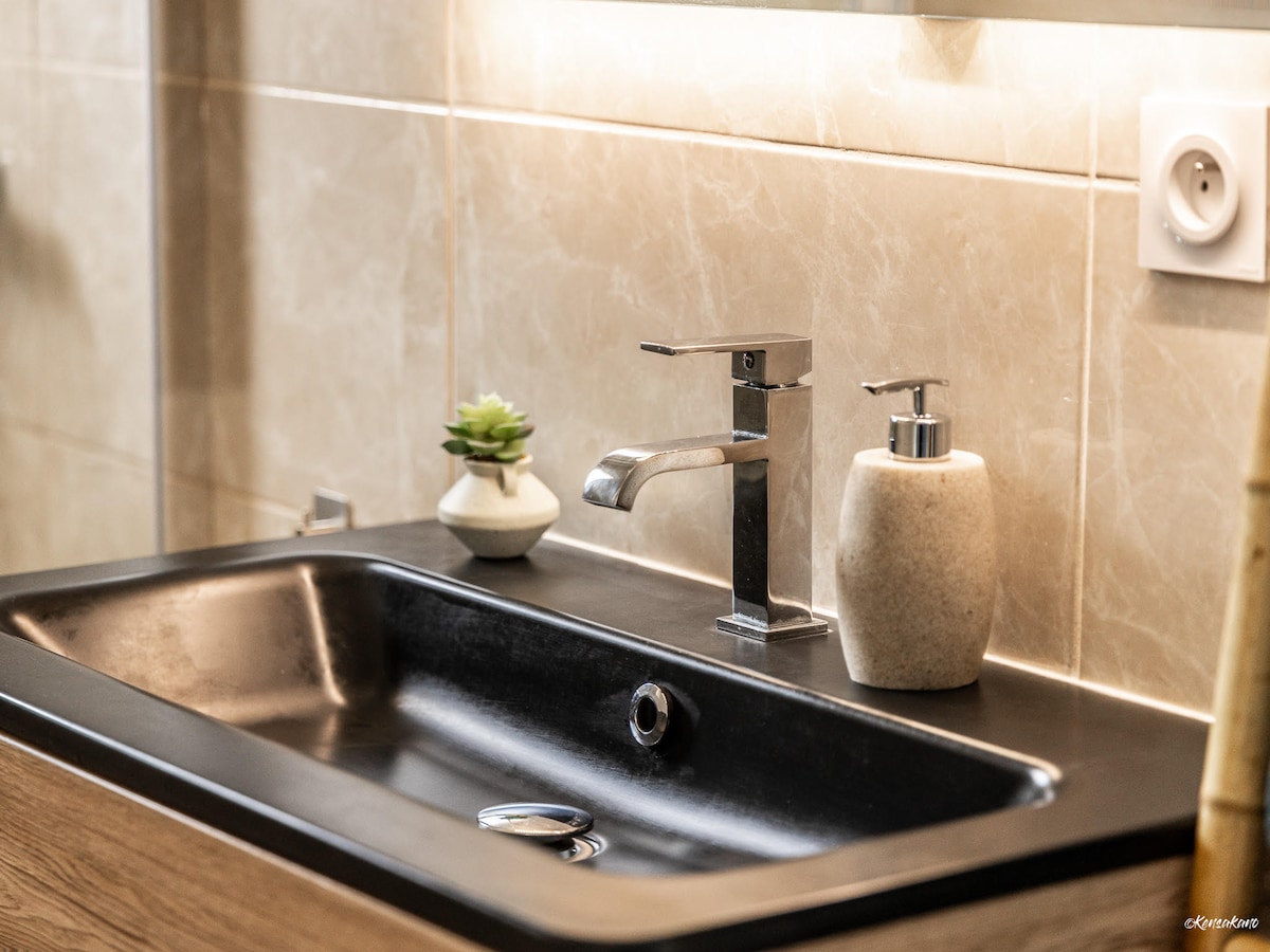 A modern bathroom sink area is featured, with a sleek black surface and a minimalistic design. A small green plant and a stone-like soap dispenser sit nearby, while warm lighting enhances the subtle textures of the tiles and fixtures.