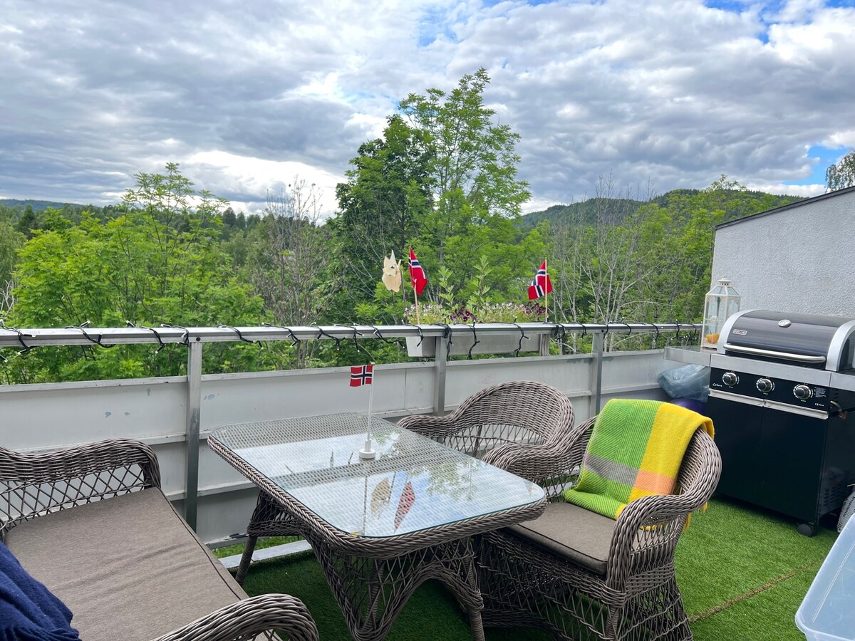 A balcony area features a glass-top table surrounded by two wicker chairs, with a colorful blanket draped over one. The space is adorned with a Norwegian flag and offers views of lush greenery, complemented by a grill located at the side.