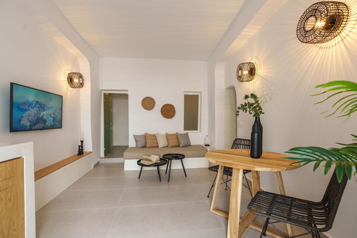 An airy living space features a neutral color palette with natural textures. A comfortable seating area with cushions is positioned against the wall, while a wooden dining table surrounded by black wire chairs provides a charming place for meals. Decorative wall accents add a touch of style.
