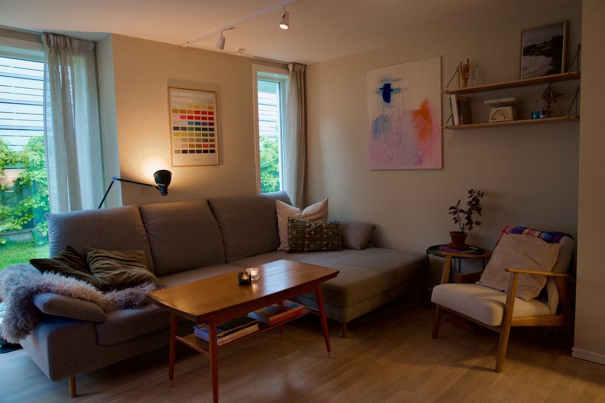A modern living area features a grey sofa with decorative pillows and a wooden coffee table at the center. Natural light enters through two windows, illuminating a colorful wall art piece and a small bookshelf with curated items. A comfortable chair adds to the inviting design.