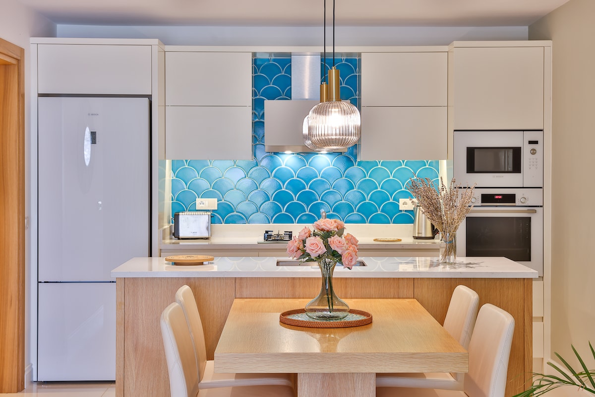 A modern kitchen displays a sleek design with white cabinetry and striking blue tiled backsplash. A dining table is set with a floral centerpiece, complemented by four light-colored chairs. Appliances include a refrigerator, oven, and microwave, showcasing a functional and aesthetically pleasing space.