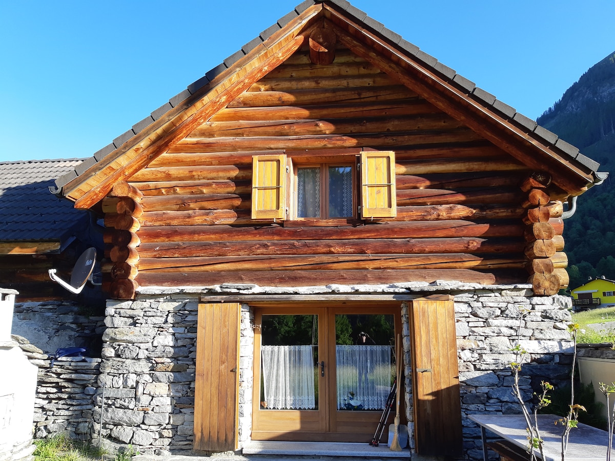 A charming mountain cabin features a rustic wooden facade with logs and stone accents. Yellow window shutters frame the upper windows, while a large double door leads into the interior. The surrounding landscape includes lush greenery and distant mountains.