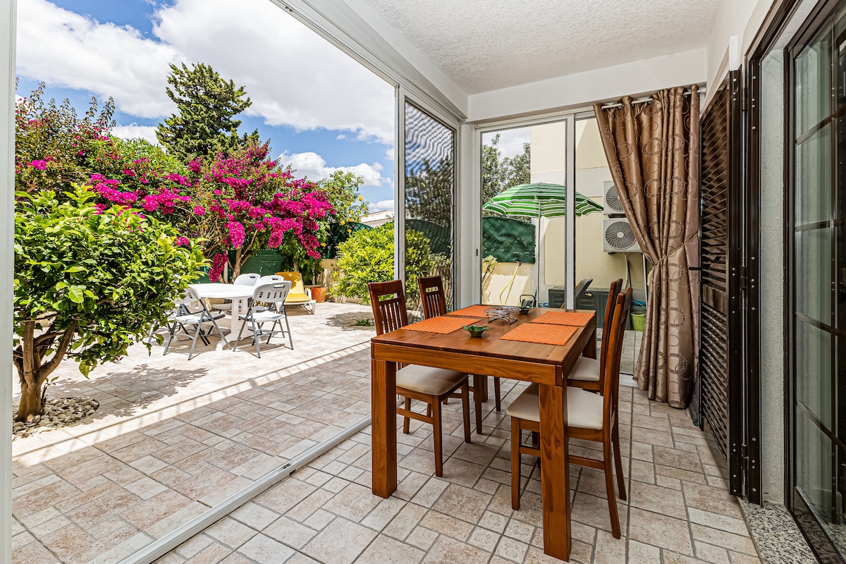 A dining area is showcased with a wooden table set for four, adjacent to a patio door that provides a bright view of a garden. Lush blooming flowers and green foliage are visible outside, along with outdoor seating options under a parasol.