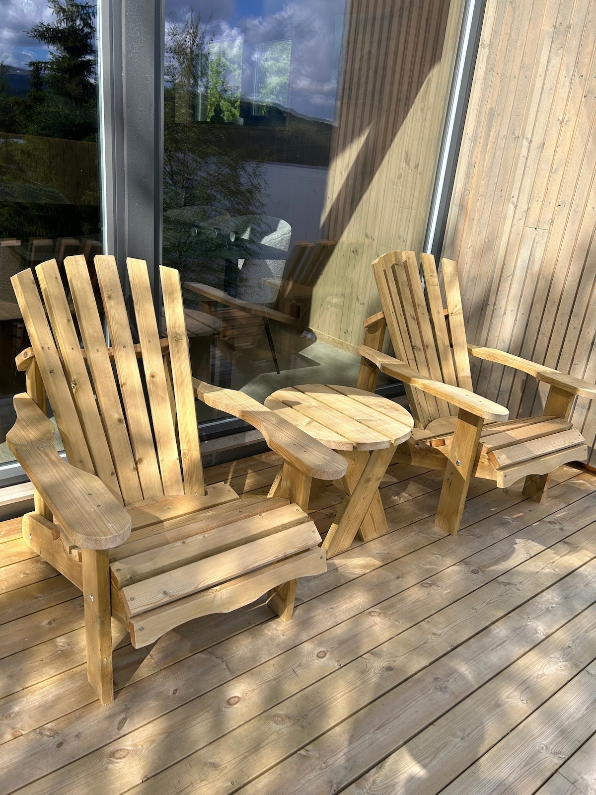 Two wooden Adirondack chairs and a small side table are arranged on a wooden deck, offering a comfortable outdoor seating area. Sunlight creates a warm glow on the natural wood, with reflections visible in the nearby glass doors.