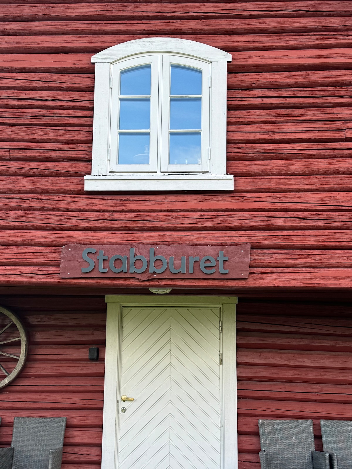 A quaint wooden facade is highlighted by a prominent sign labeled 'Stabburet'. A white door features a diagonal panel design, framed by a rustic red exterior. A charming window above allows natural light to enhance the inviting entrance. Two seating arrangements are visible beneath the window.