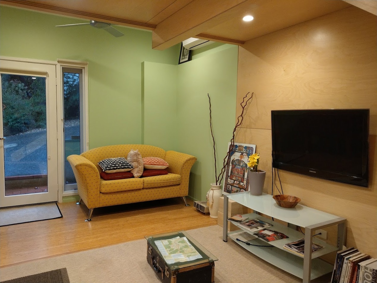 A lounge area is presented with light green walls and wooden finishes. A yellow sofa with patterned cushions is positioned against the wall, accompanied by a small coffee table displaying magazines. A flat-screen TV is mounted nearby, and natural light is allowed in through large glass doors.