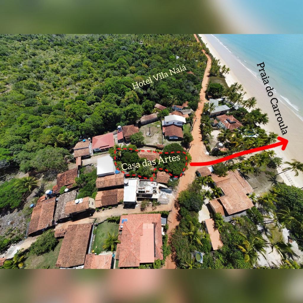 An aerial view captures the layout of Casa das Artes nestled within a lush green forest, with the coastline of Praia do Carroula visible nearby. The image shows surrounding structures and a sandy path leading to the beach, emphasizing the tranquil and natural setting.