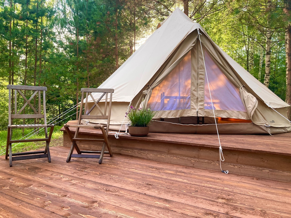 A spacious glamping tent is set on a wooden terrace, surrounded by trees. Two wooden chairs are positioned nearby. The tent's entrance is open, allowing natural light to illuminate the interior, showcasing a serene and inviting environment.