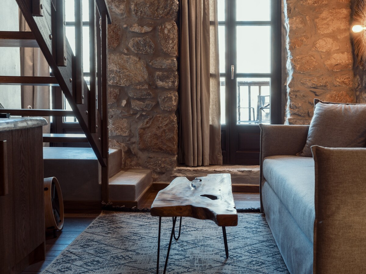 A cozy living area features a light grey sofa alongside a unique wooden coffee table, showcasing natural grain patterns. A stone wall adds texture to the space, while large glass doors provide views and natural light. Soft curtains frame the windows, enhancing the serene ambiance.
