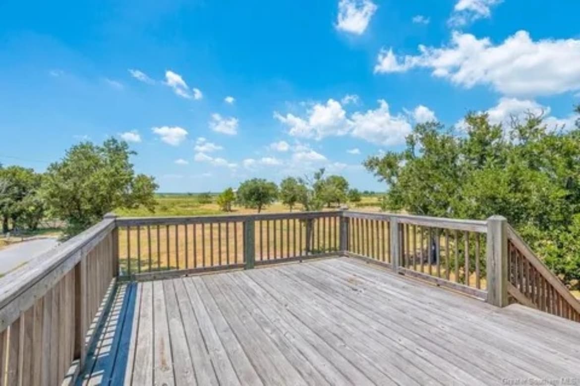 A deck with wooden railing offers an unobstructed view of the surrounding landscape, featuring green fields and scattered trees under a bright blue sky with scattered clouds. The sunlit deck invites relaxation and enjoyment of the outdoor scenery.