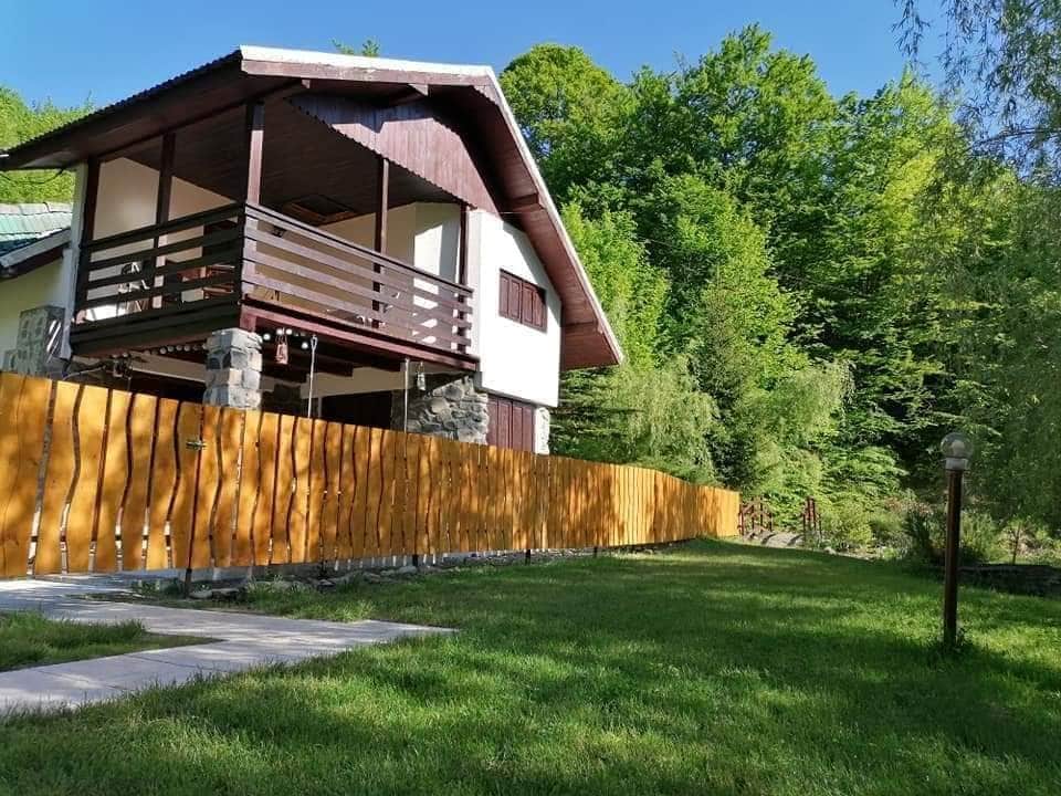 A two-story house is surrounded by lush greenery, featuring a wooden balcony that overlooks a spacious yard. A neatly maintained wooden fence lines the property, complementing the natural environment. Pathways are visible leading towards the entrance, set against a backdrop of trees.