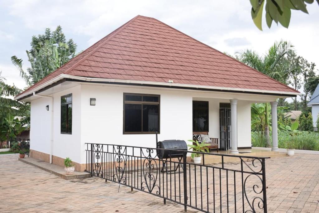 The exterior of a single-story home is depicted, featuring a unique gabled roof and white walls. A covered porch is visible, along with a railing outlining the entrance. Lush green trees and plants surround the property, contributing to the tranquil environment.