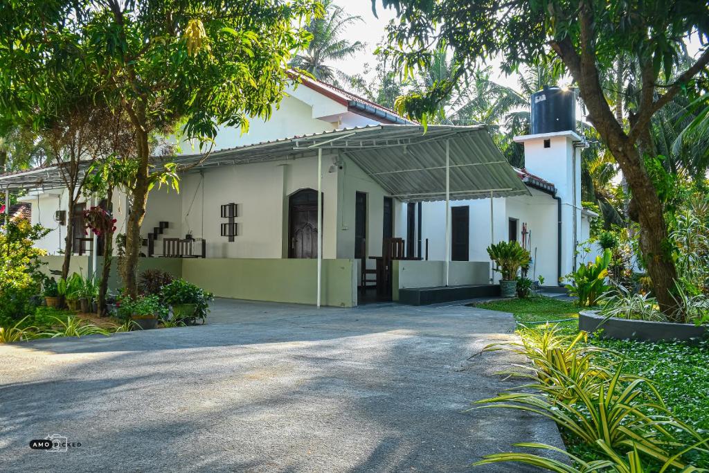 The exterior of the villa is featured, showcasing a broad covered porch and a spacious driveway. Lush greenery surrounds the property, with tropical plants and trees providing shade. The building has a clean, white façade with contrasting decorative elements visible.