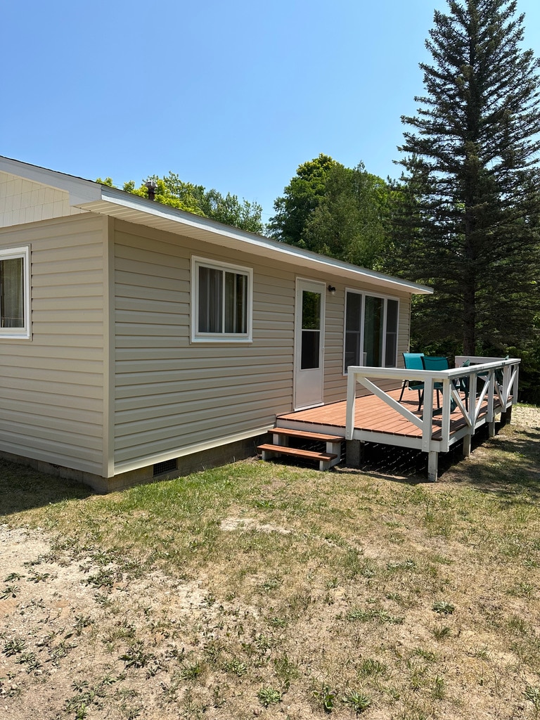The exterior of the cottage is showcased with light-colored siding and multiple windows. A wooden deck with white railings provides access to the entrance, which features a glass sliding door. Surrounding greenery and a tree are visible in the background.