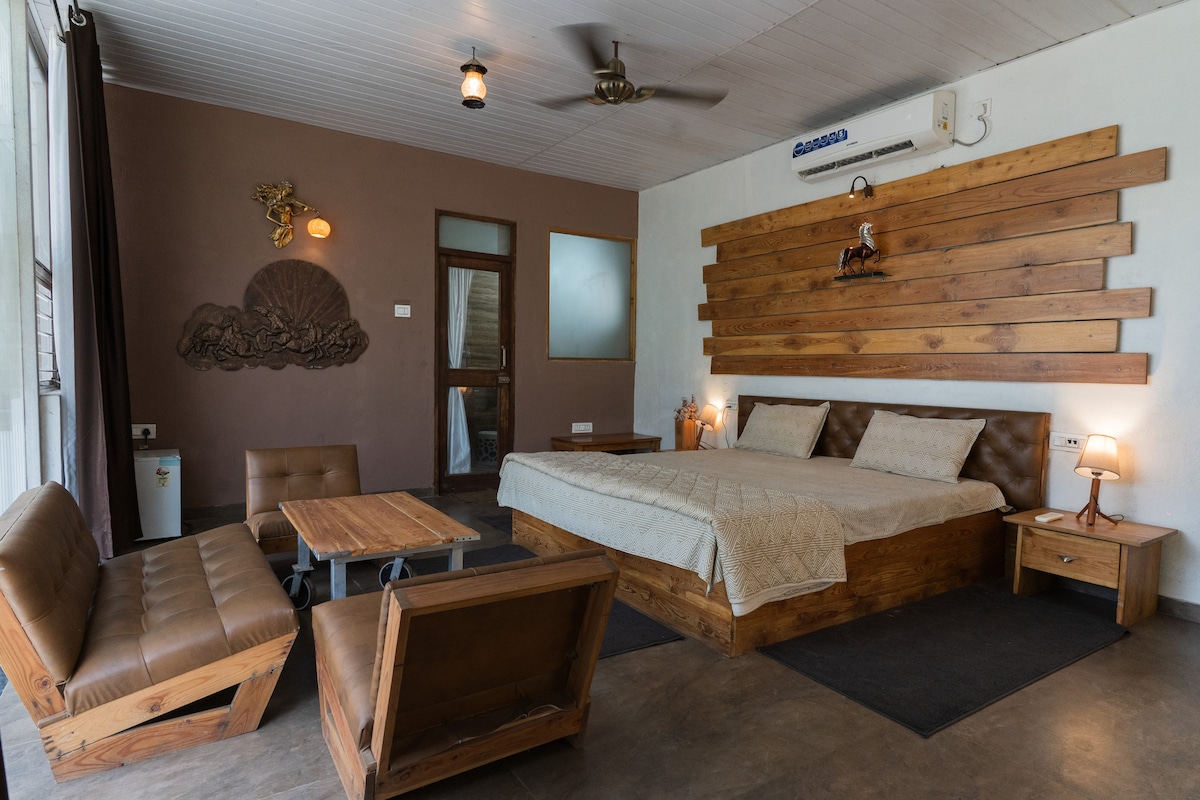 An inviting bedroom features a spacious wooden bed with a light-colored quilt, complemented by wooden accents on the walls. Two seating areas, a small table, and warm lighting create a cozy atmosphere. A window allows natural light to enter, enhancing the room's overall appeal.