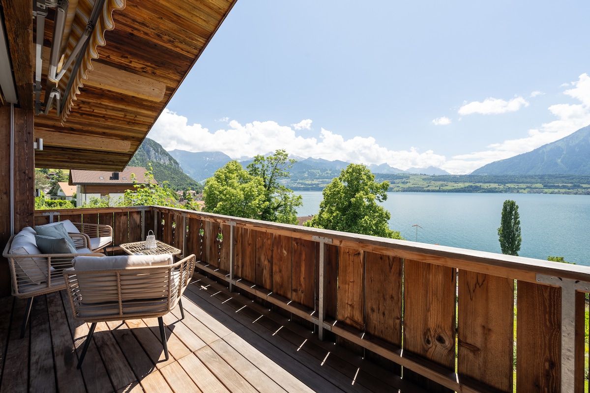 A spacious balcony offers expansive views of the Thunersee and surrounding mountains. Outdoor seating arrangements create a place for relaxation, with wooden railings framing the picturesque landscape. The natural light highlights the wooden flooring, contributing to a welcoming atmosphere.