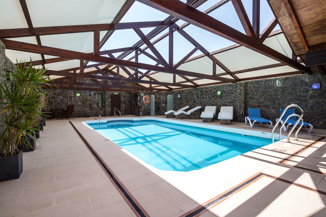 An indoor swimming pool is framed by a wooden structure with clear roofing, allowing natural light to fill the space. Sun loungers are arranged alongside the pool, while lush potted plants enhance the serene environment.
