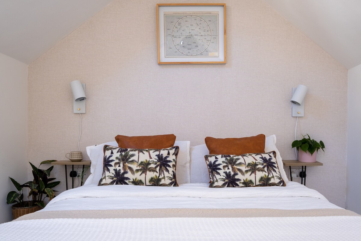 A cozy mezzanine sleeping area is showcased, featuring a double bed adorned with soft linens and two decorative pillows with a palm tree motif. Wall-mounted lamps are positioned on either side of the bed, complemented by a small plant and framed artwork above.