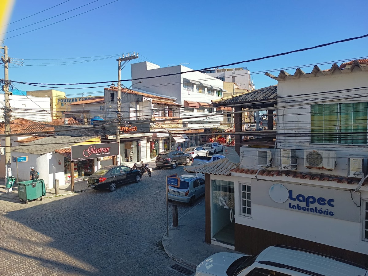A bustling street scene is captured, showcasing a mix of local shops and parked vehicles. Cobblestone streets lead through the center of the area, bordered by buildings with colorful façades. Power lines are visible overhead against a clear blue sky, reflecting a lively urban atmosphere.