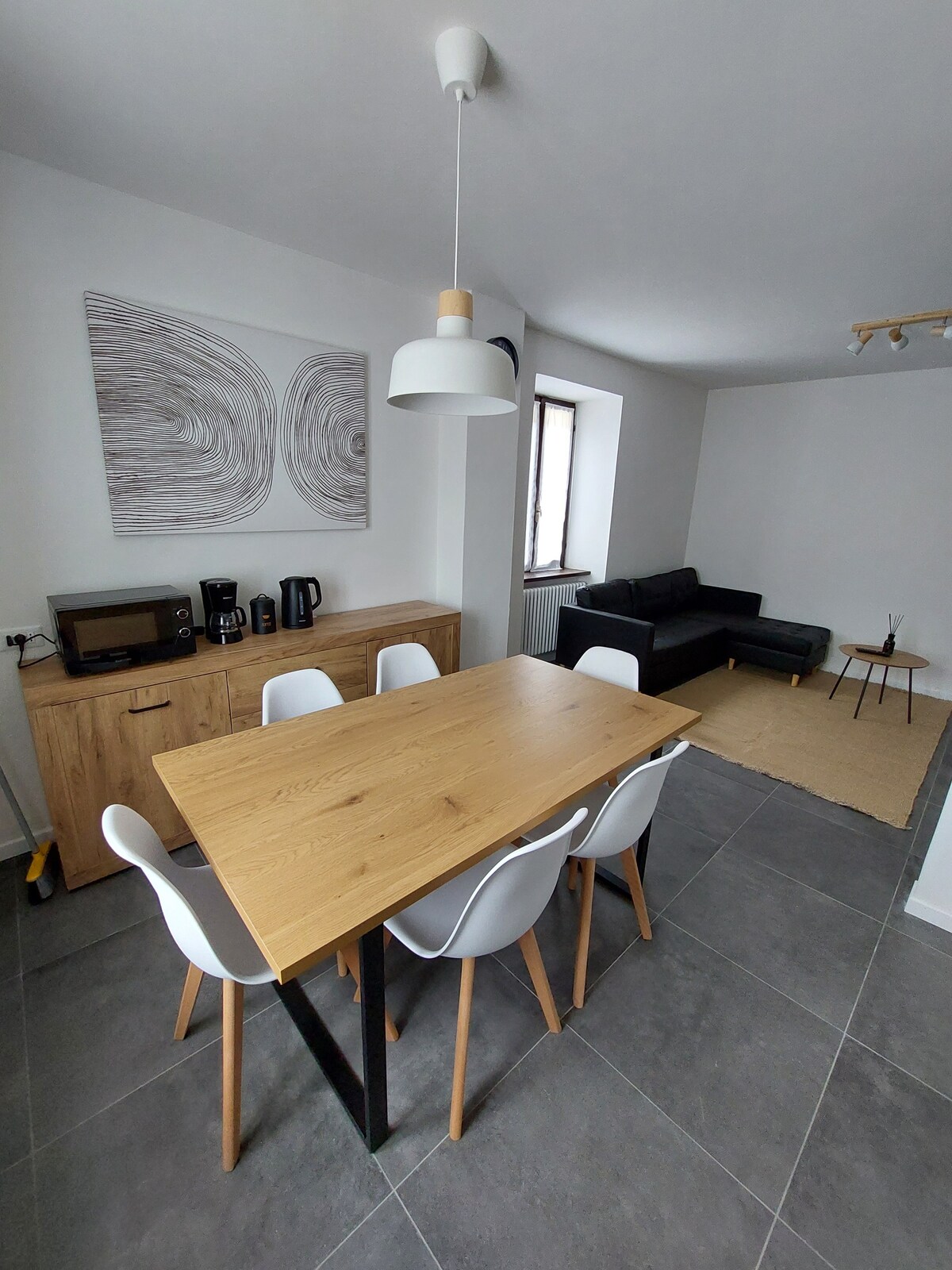 A bright dining area features a wooden table surrounded by six white chairs. A modern white pendant light hangs above the table. In the background, a dark sofa is positioned next to a subtle coffee table, with artwork on the wall enhancing the contemporary setting.