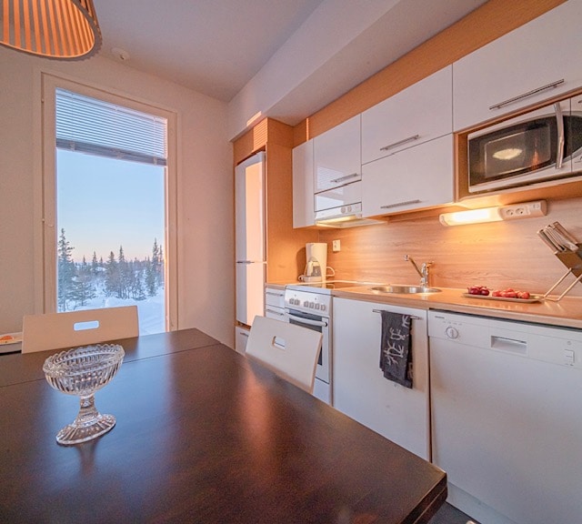 The kitchen area features modern cabinetry in warm tones, complemented by a dark wooden dining table. A large window offers views of the snow-covered landscape outside. Appliances, including a stove, microwave, and dishwasher, are integrated into the design, ensuring functionality and convenience.