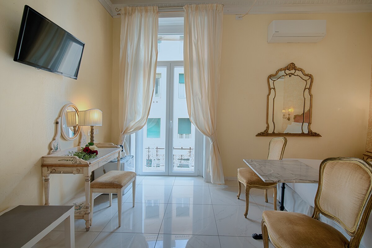 The room features a marble floor, a large window draped with light curtains, and a view of the outside. A flat-screen TV is mounted on the wall. A small table and two elegant chairs are positioned near a vanity with decorative lamps and a mirror.