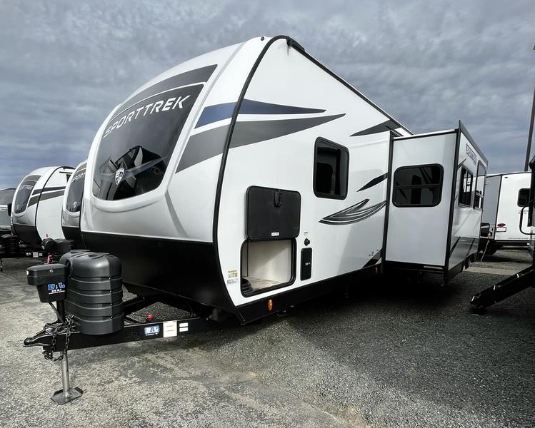 A modern travel trailer is displayed from an exterior angle, featuring a sleek white and gray design. The spacious slide-out section extends from the side, enhancing the living area inside. A black hitch is attached to the front, with outdoor storage compartments visible.