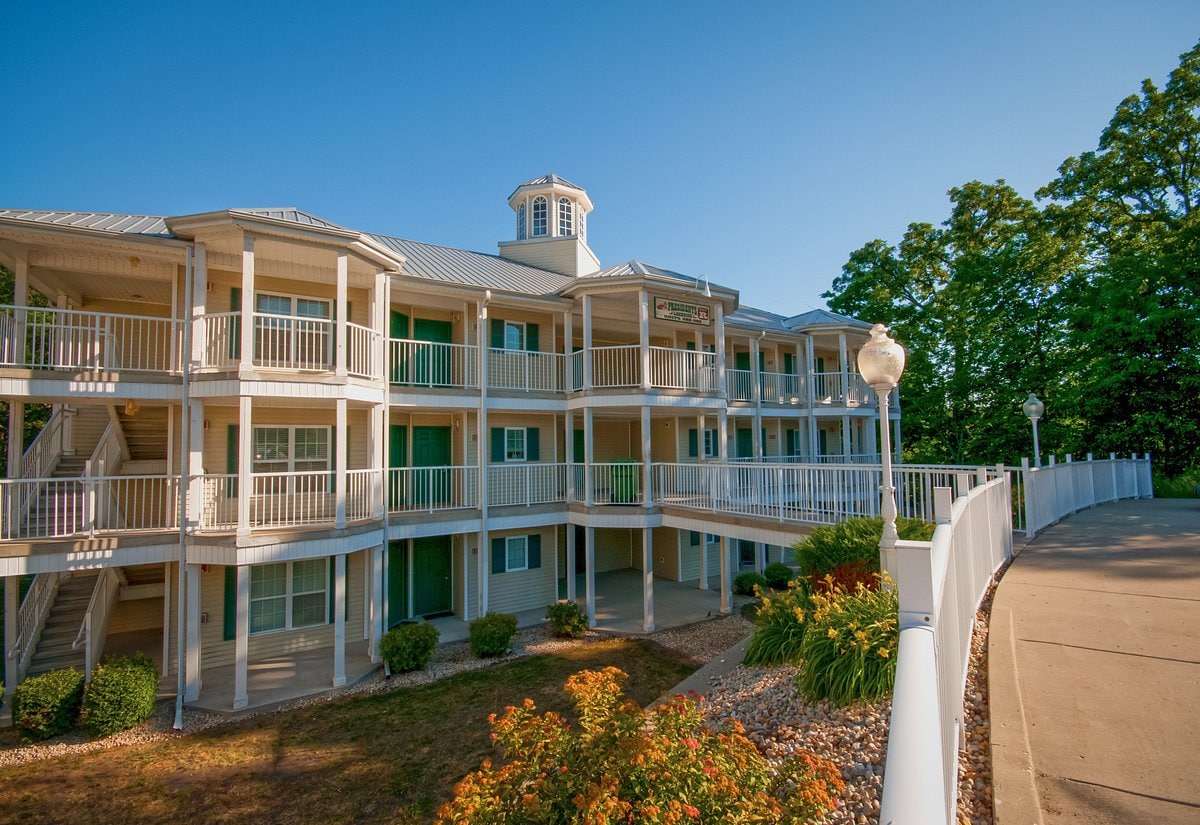 The three-story building features multiple balconies, allowing for outdoor space on each level. A well-maintained garden with colorful flowers and shrubs borders the pathway leading up to the entrance. The structure is surrounded by trees, providing a green backdrop.