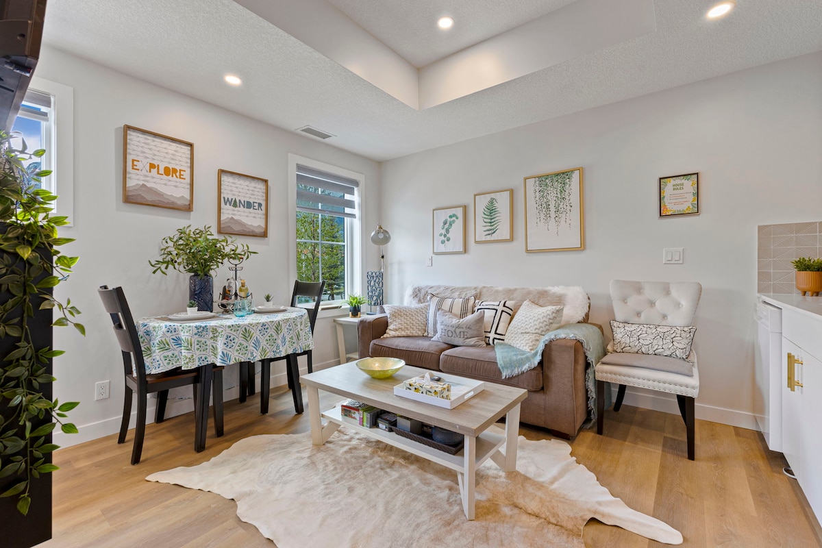 The bright living area features a modern sofa accompanied by an armchair, a coffee table, and a dining table set for four. Natural light enters through multiple windows, enhancing the space adorned with tasteful decor and greenery that adds a fresh touch.