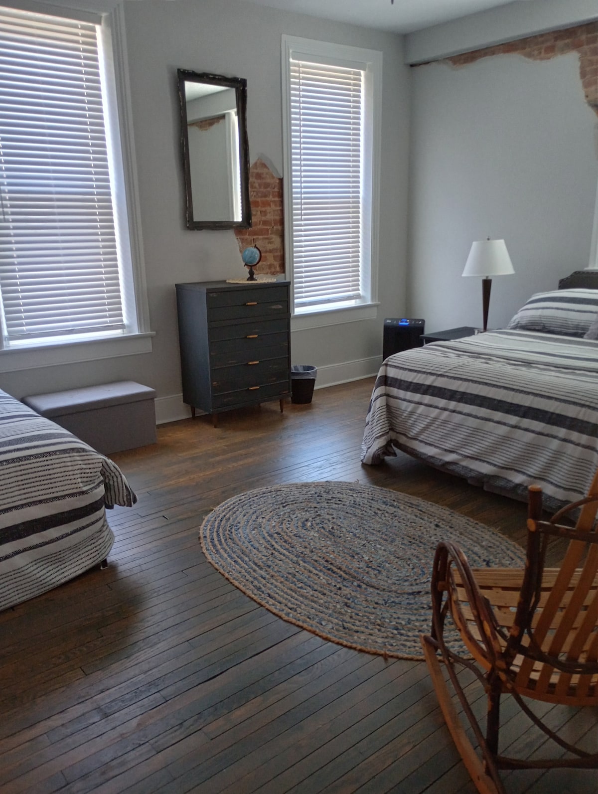 A spacious bedroom is furnished with a queen bed and a single bed, both dressed in striped linens. Natural light filters through the two vertical-closed windows. A small dresser and a lamp are positioned nearby, enhancing functionality. A circular area rug rests on the hardwood floor.