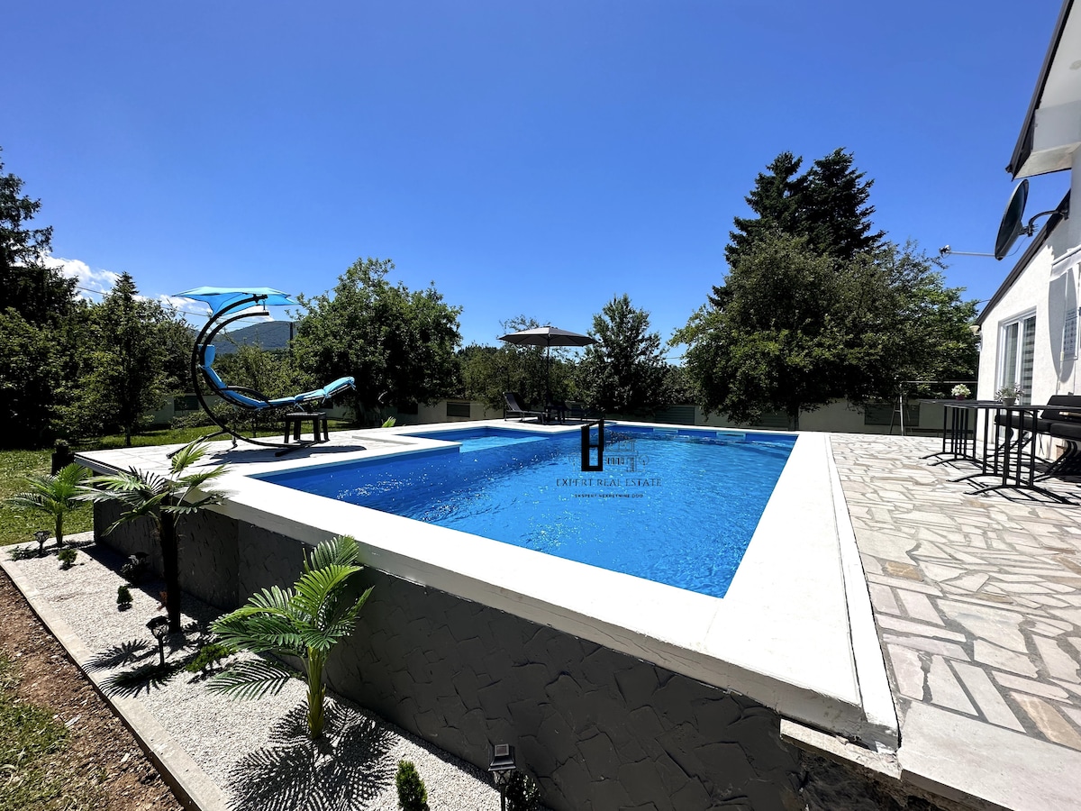 A serene pool area features a large rectangular pool surrounded by a stone patio. Lush greenery complements the space, including palm plants. A comfortable lounge chair sits beside the pool, inviting relaxation under a clear blue sky.