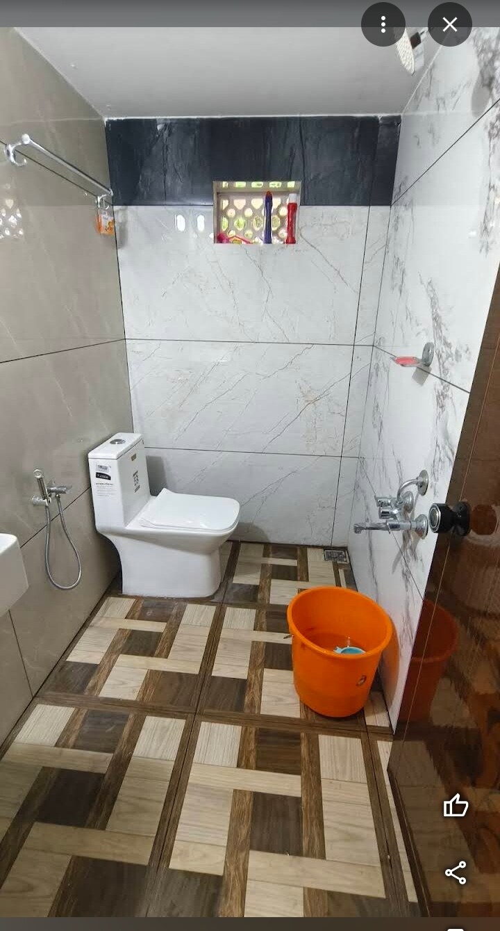 The bathroom features modern tile flooring with a geometric pattern. A white toilet and a wall-mounted sink are present, along with a shower area equipped with a chrome faucet. A bright orange bucket adds a pop of color, and natural light filters through a small window.