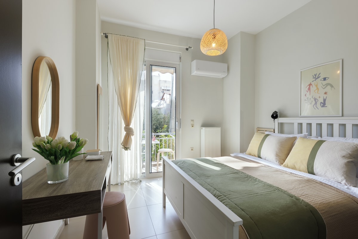 A bright bedroom features a spacious bed with neutral-toned linens and decorative pillows. A large window with sheer curtains allows natural light, while a small desk and a mirror provide functional elements. A pendant light hangs from the ceiling, adding a touch of warmth.