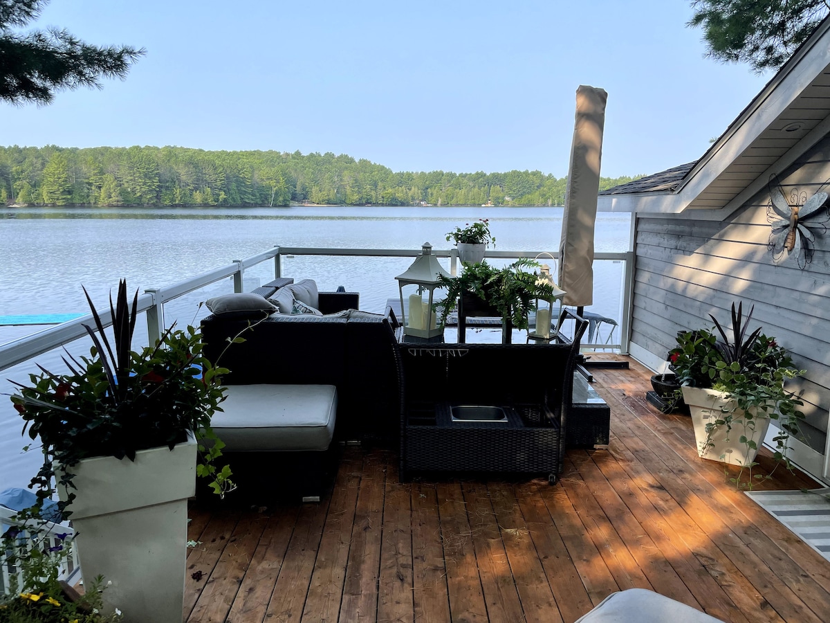 A spacious deck overlooks the tranquil lake, adorned with comfortable seating arrangements and potted plants. Sunlight casts gentle shadows on the wooden floor, enhancing the serene outdoor setting. A tall umbrella provides shade for relaxation beside the water.
