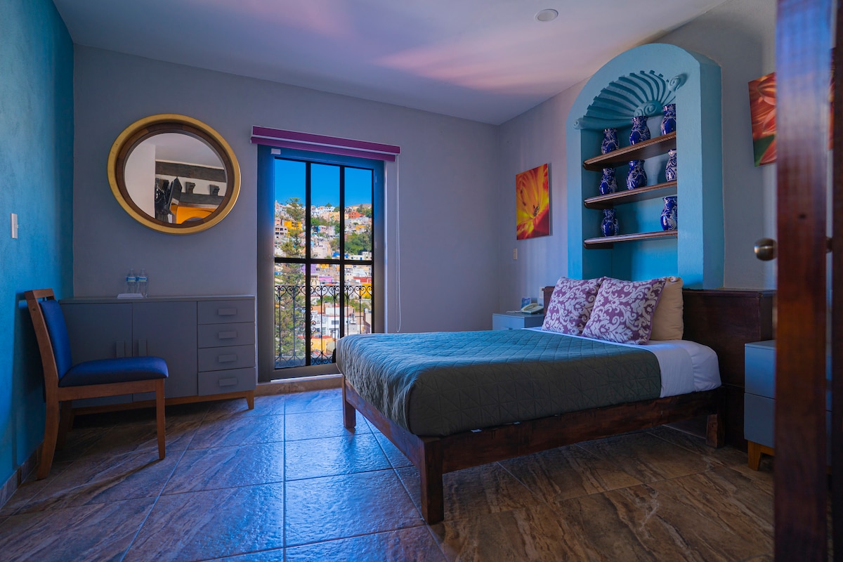 A bright bedroom features a spacious bed with a patterned coverlet. A round mirror hangs above a decorative shelf adorned with pottery. Natural light filters through a window, revealing views of the city. A wooden chair and a nightstand add to the room's functionality.