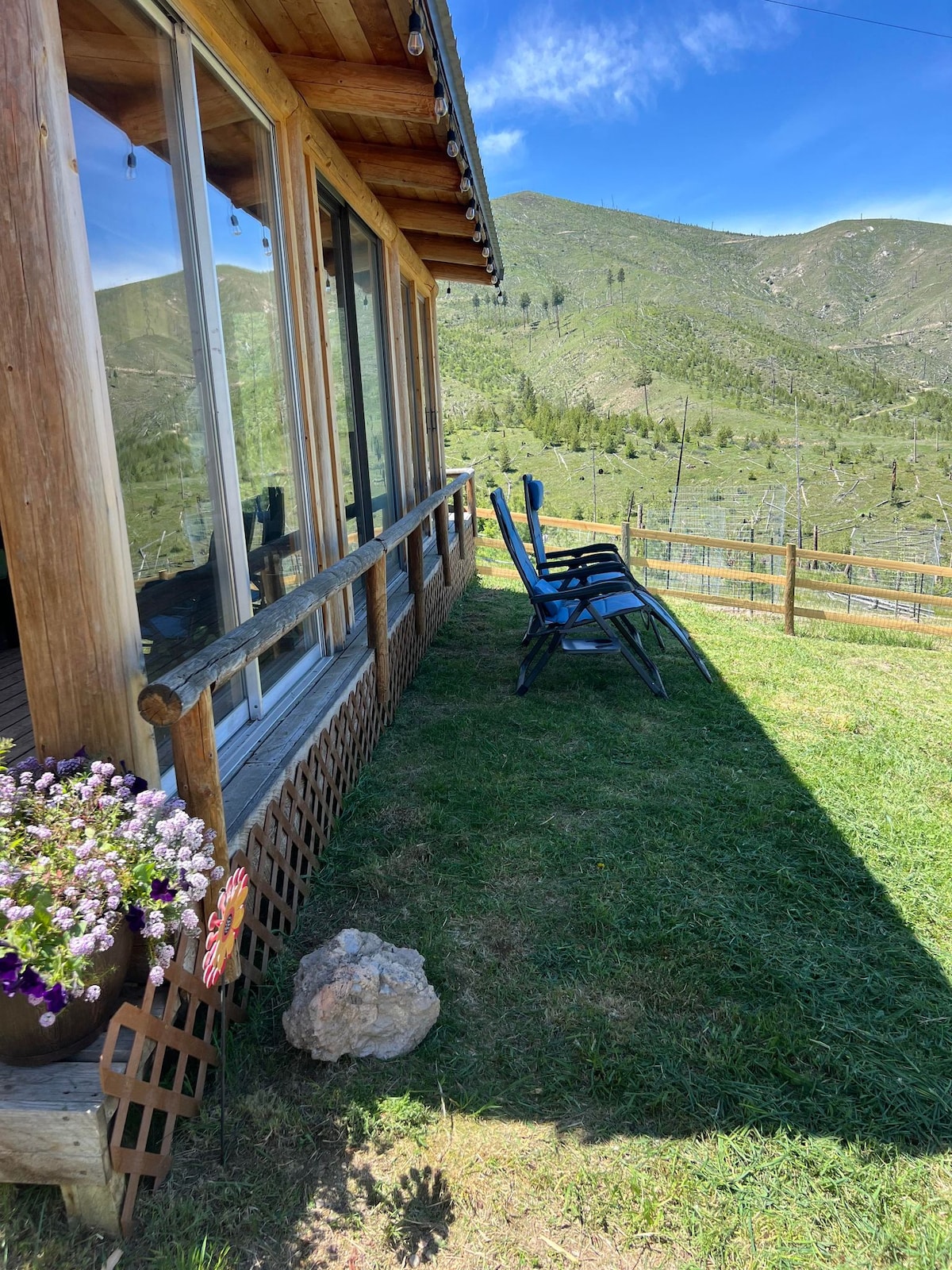 The exterior of the cabin showcases a wooden deck with two chairs inviting relaxation. A flower pot with vibrant blooms sits near the railing, while grassy areas lead toward scenic hills in the background, partially shadowed by the cabin's overhang.